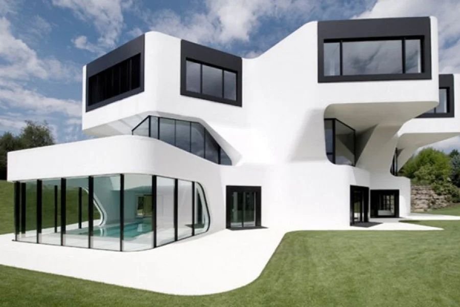 Geniş yeşil bir çim alan üzerinde yer alan, organik kıvrımlı hatlara, siyah pencere çerçevelerine ve zemin katta kapalı cam bölmelere sahip ultra modern beyaz lüks villa mimarisi.