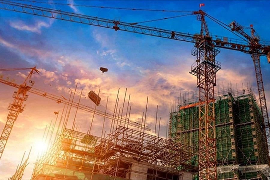 Gün batımında, çok sayıda kule vincin aktif olarak çalıştığı ve güvenlik ağları ile iskele sistemleriyle çevrili yüksek bir binanın yer aldığı büyük ölçekli, modern inşaat şantiyesi genel görünümü.